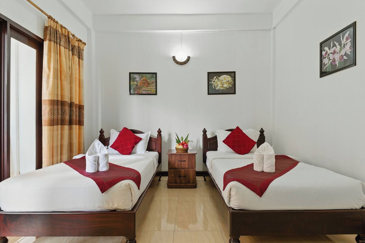 Habitación de hotel con dos camas, ropa de cama roja y blanca, una mesita de noche de madera con una planta y obras de arte en las paredes.