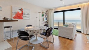 A modern living room with a round table, bar stools, a sofa, and a large window offering a view of the beach. - 1 bedroom accommodation in De Panne (De Panne)