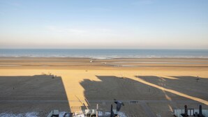 A wide beach view with a clear sky, shadows of objects on the sand, and a few people in the distance. - 1 bedroom accommodation in De Panne (De Panne)