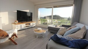 A modern living room with a flat-screen TV, a sofa, a coffee table, and a view of a green landscape through large windows.
