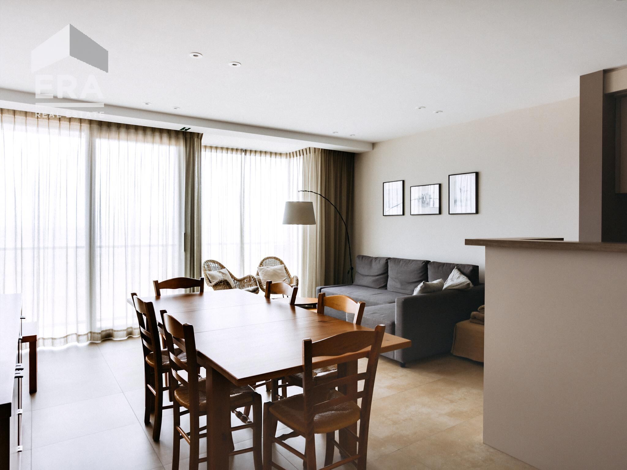 A modern living room with a dining table and chairs, a grey sofa, and a large window with curtains.