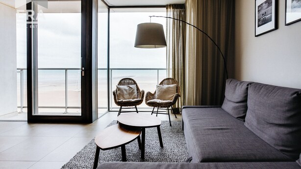 A modern living room with a grey sofa, two wicker chairs, a small table, and a floor lamp. A large window offers a view of the beach. - 3 bedroom accommodation in Koksijde (Koksijde)