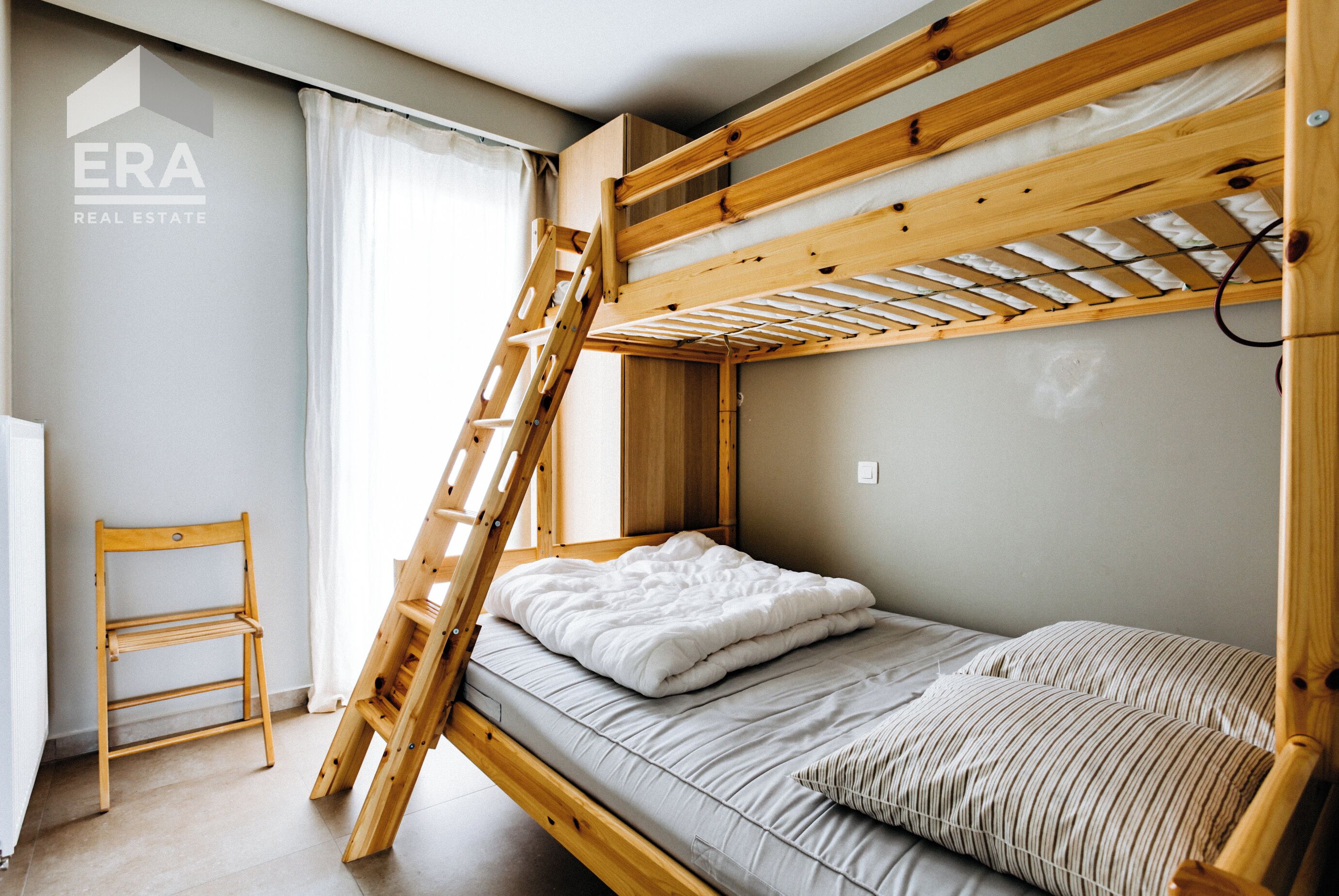 Una cama litera con una escalera de madera, una cama individual con colchón blanco y almohada a rayas, y una silla de madera.
