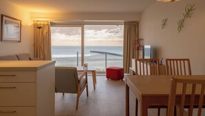 A room with a large window overlooking the beach, a dining area with wooden furniture, and a television on a stand. - 1 bedroom accommodation in Nieuwpoort (Nieuwpoort)