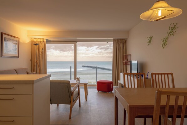 A room with a large window overlooking the beach, a dining area with wooden furniture, and a television on a stand.