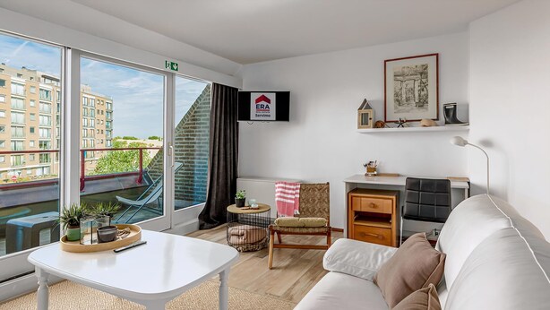 A modern living room with a white sofa, a small table with plants, a chair, a TV mounted on the wall, and a view of a building outside. - 2 bedroom accommodation in Koksijde (Koksijde)