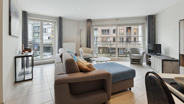 Un soggiorno moderno con un divano, un tavolo da pranzo e una TV. È presente un balcone con vista sugli edifici.