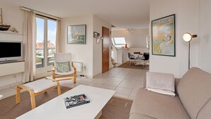 A modern living room with a flat-screen TV, a white coffee table with a book, a beige sofa, and a dining area in the background. - ZeeZonZand C-0206 + G* (Koksijde)