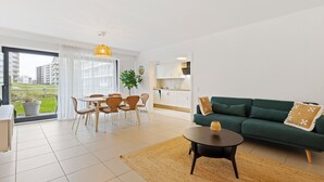 A modern living room with a green sofa, a dining table with chairs, a kitchen in the background, and a large window with a view of buildings. - 1 bedroom accommodation in De Panne (De Panne)