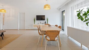 A modern dining area with a wooden table and wicker chairs, a flat-screen TV mounted on the wall, and large windows with sheer curtains. - 1 bedroom accommodation in De Panne (De Panne)