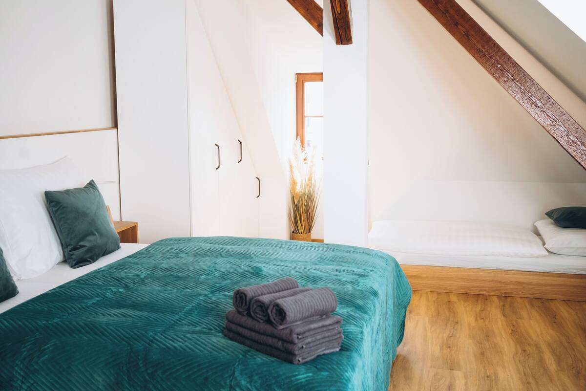 Une chambre avec une couvre-lit turquoise, un lit soigneusement fait avec des serviettes pliées, une tête de lit en bois, un plancher en bois et une fenêtre donnant sur l’extérieur.