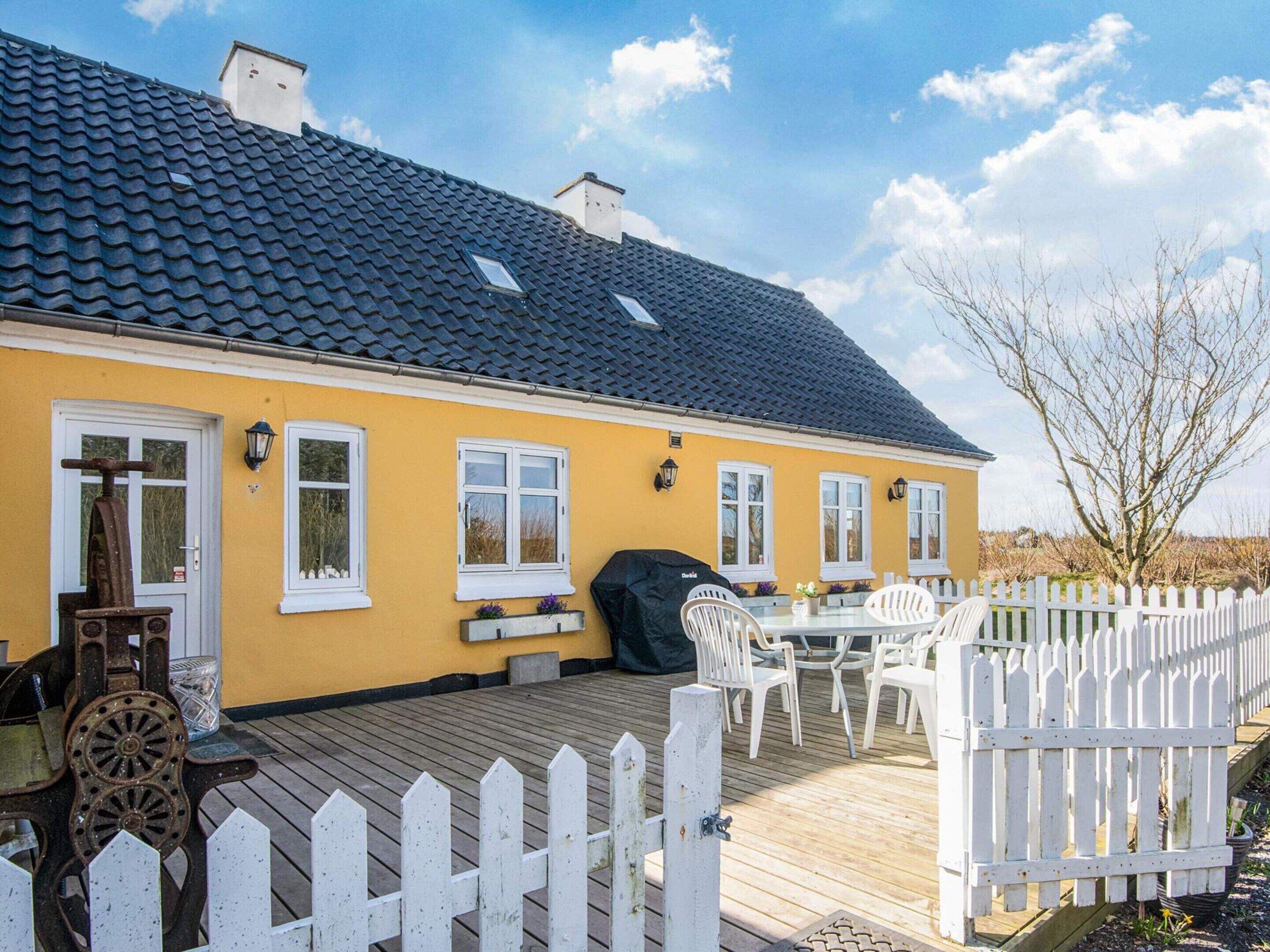 Una casa amarilla con techo oscuro, cerca blanca en forma de piquete y un porche con mobiliario exterior.