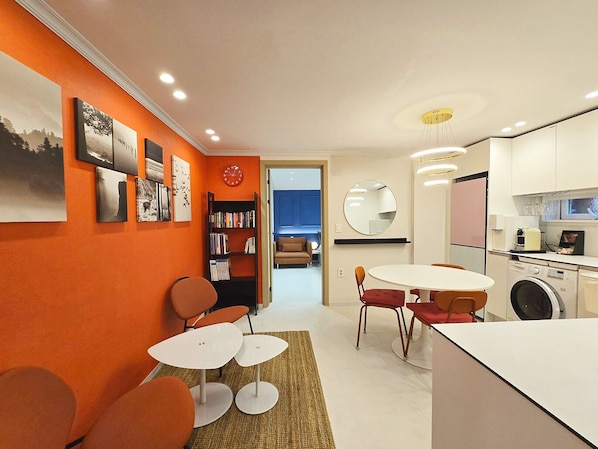 A modern kitchen with white cabinets, a washing machine, and a dining area with a round table and orange chairs.