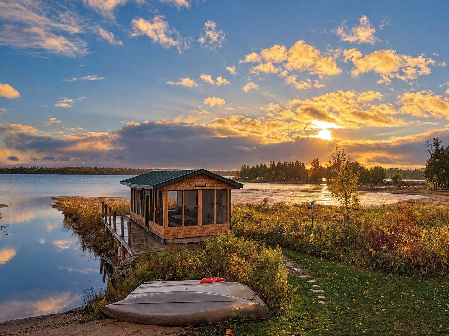 A wooden cabin with a glass enclosure, situated by a calm lake with a dock extending into the water.