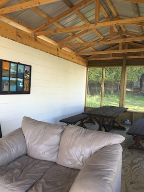 Een overdekt terras met een houten bank, een bank met kussens, een picknicktafel en een muur met een ingelijst beeld.