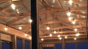 An outdoor seating area with wooden tables and benches, large windows, and string lights on the ceiling. - The Westender Retreat | Amazing Lake Views & Waterfront Dock House (Hessel)