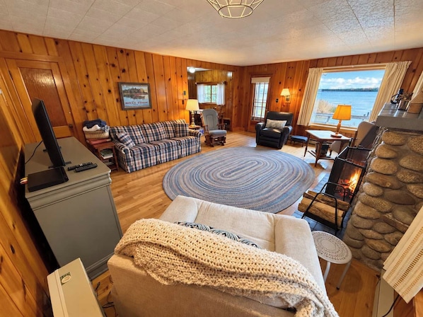 Un intérieur de chalet chaleureux avec une cheminée, un canapé, un téléviseur et une vue sur l’extérieur.