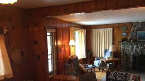A wooden cabin interior with a dining area, a living room with a stone fireplace, and a view of the outdoors through a window. - The Westender Retreat | Amazing Lake Views & Waterfront Dock House (Hessel)