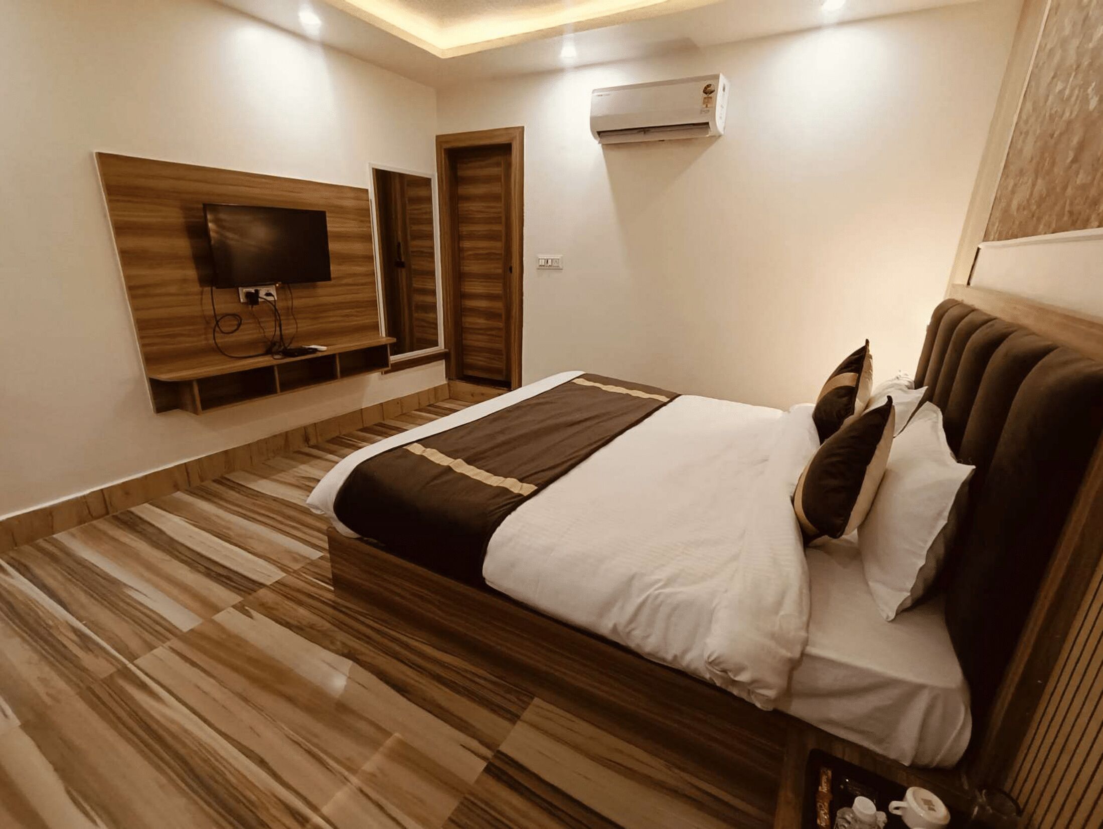 A modern bedroom with a large bed, wooden flooring, a mounted TV, and an air conditioning unit.