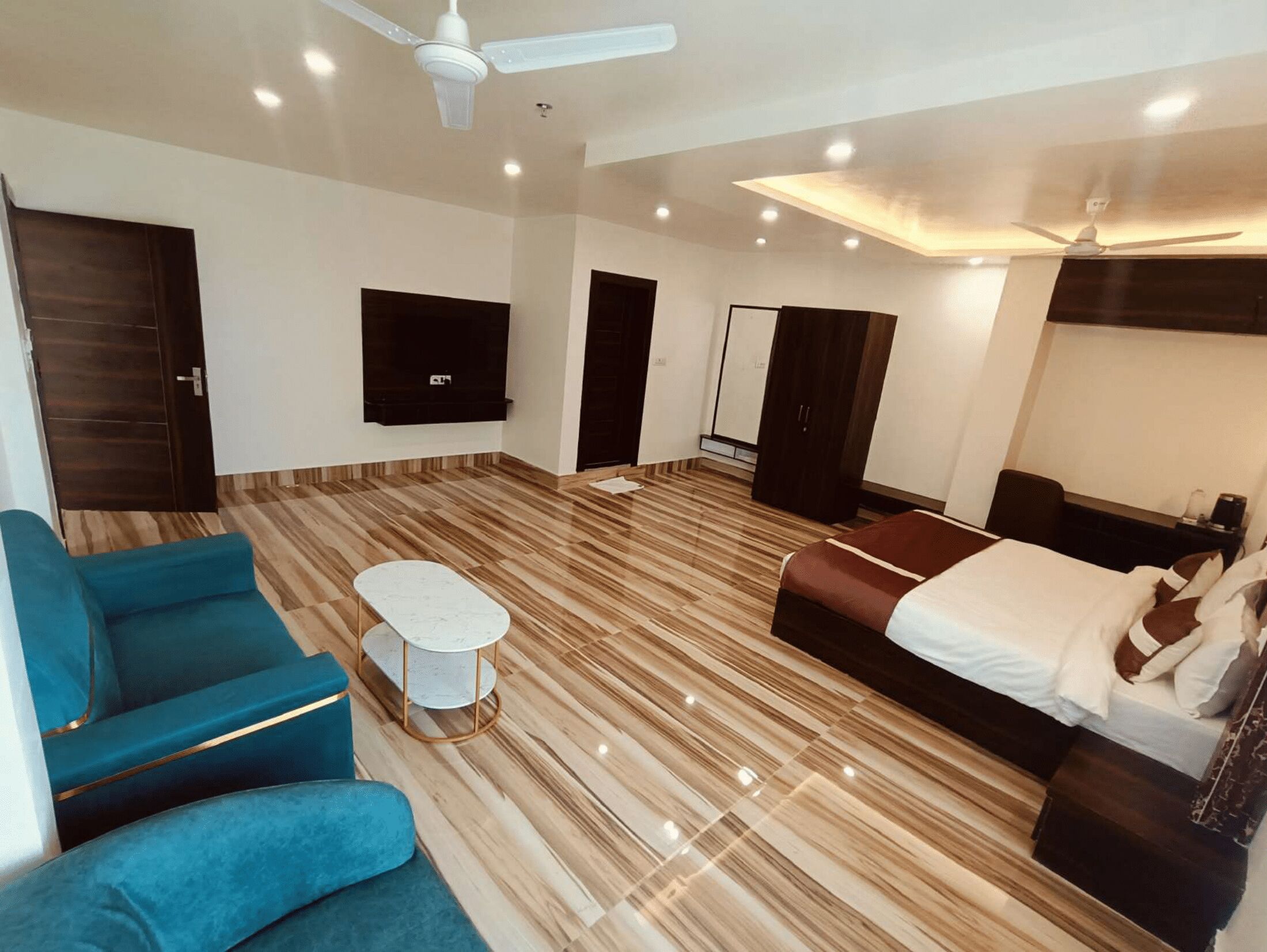 Une chambre d’hôtel moderne avec un grand lit, deux canapés bleus, une petite table ronde, un téléviseur fixé au mur et un ventilateur de plafond.
