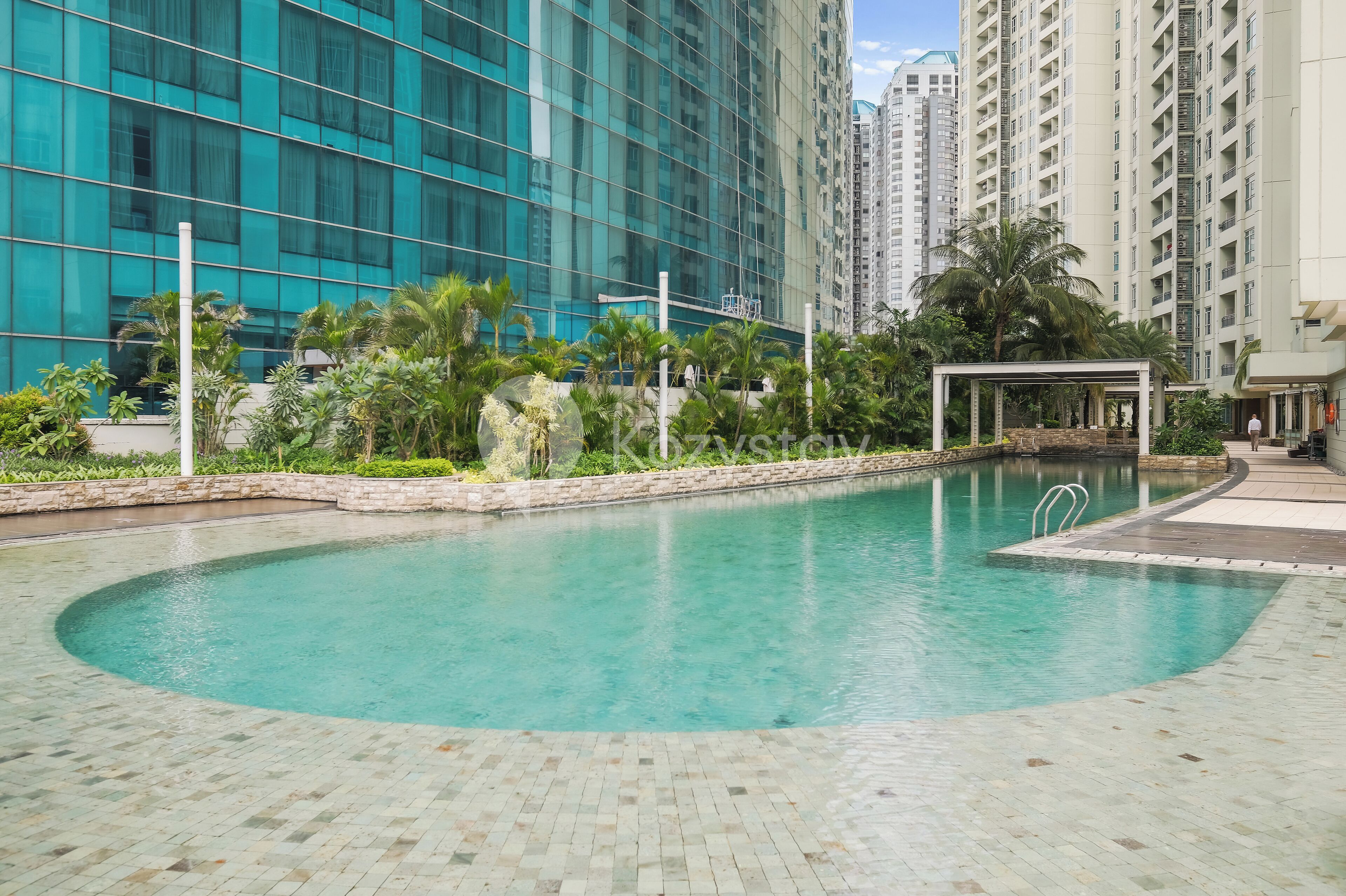 A swimming pool with a tiled deck, surrounded by greenery and tall buildings.