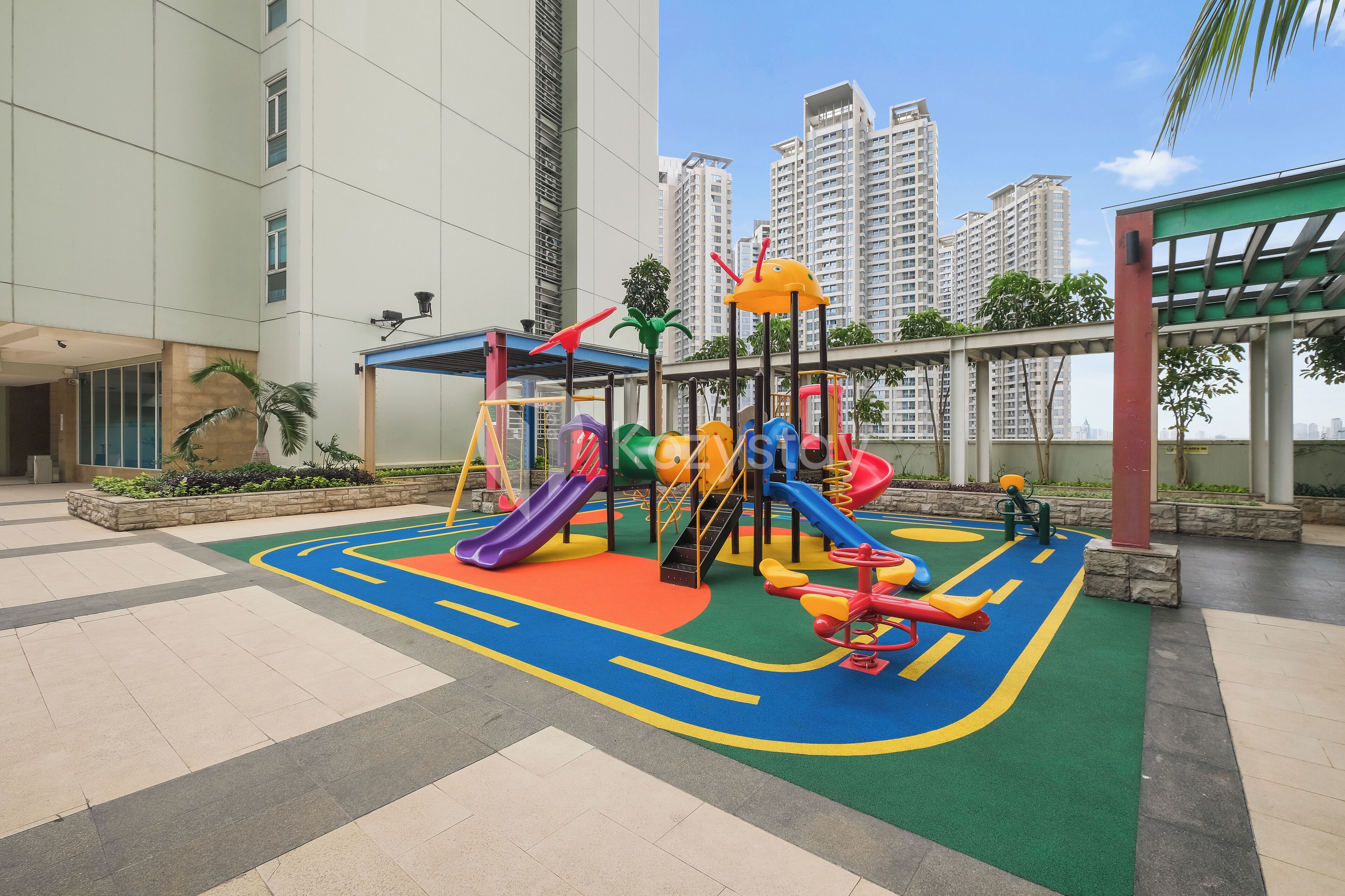An outdoor playground with slides and climbing structures, surrounded by a paved area and tall residential buildings in the background.