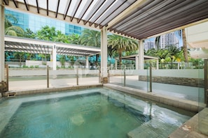 Un espace piscine doté de rampes en verre, d’un coin salon couvert et offrant une vue sur les palmiers et les bâtiments.