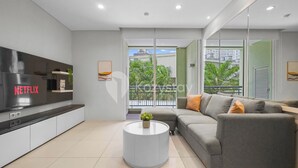 Een moderne woonkamer met een flatscreen-tv, een grijze bank, een ronde salontafel en een balkon met uitzicht op gebouwen en groen.