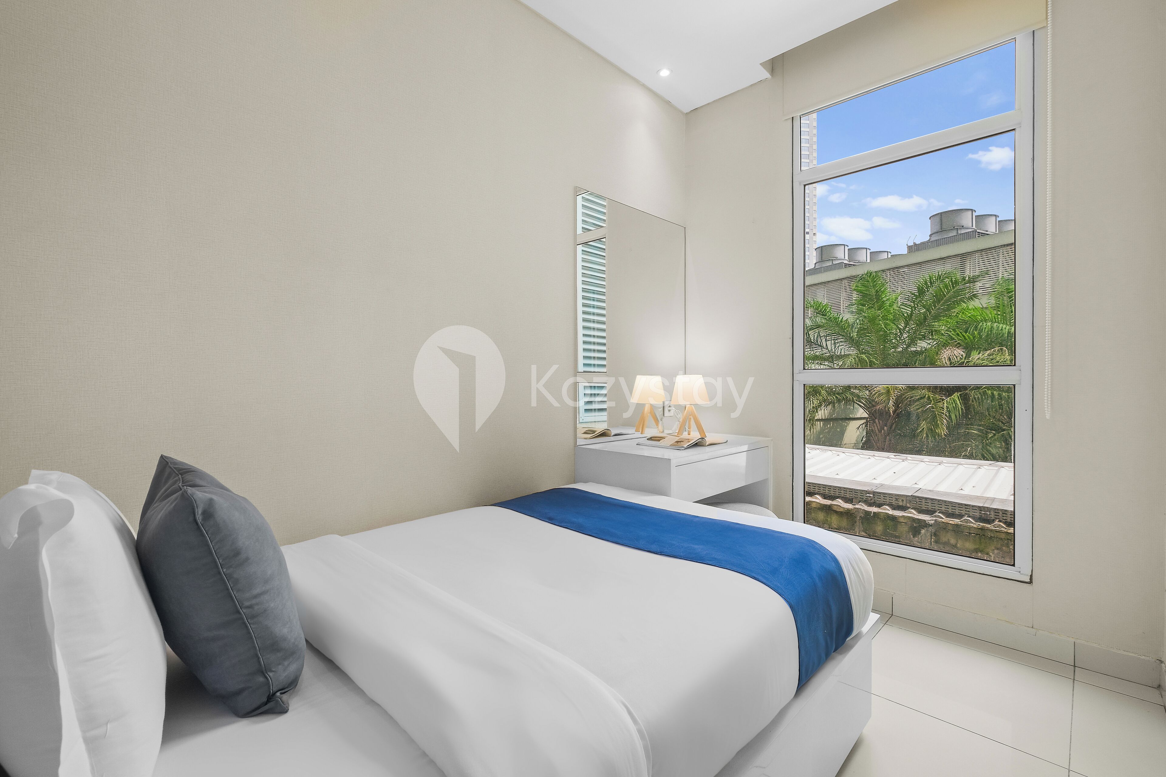 A modern bedroom with a bed, a nightstand, a mirror, and a large window offering a view of buildings and greenery.