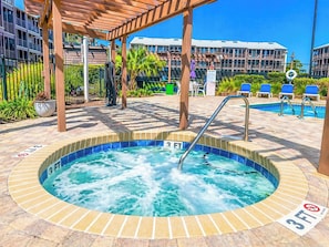 A hot tub with a wooden pergola overhead, surrounded by lounge chairs and a pool.