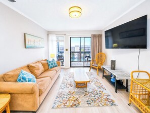 A living room with a sofa, a coffee table, a TV, and a view through a glass door. - Tilghman Beach and Racquet Club 358 (North Myrtle Beach)