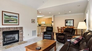 A living room with a fireplace, leather sofas, a dining area with wooden chairs, and a kitchen in the background. - Mountain View Fore You - Golf Front 2 BR, 2BA Condo (Maggie Valley)