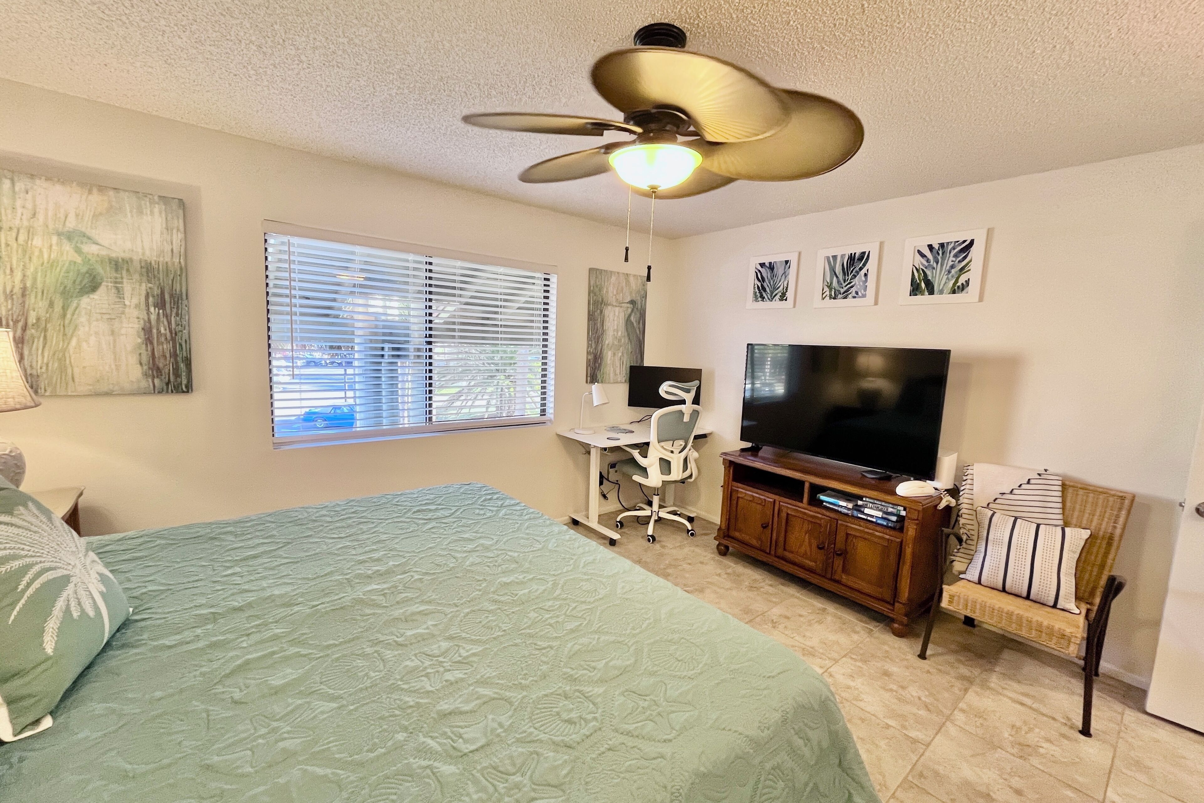 A bedroom with a bed, a desk, a chair, a television, and a ceiling fan.