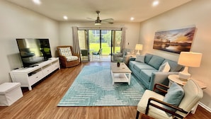 A living room with a TV, sofa, chairs, and a view through a glass door to a patio. - Best Address at the Beach, Gold-rated condo just a short walk to the beach! (Saint Augustine)