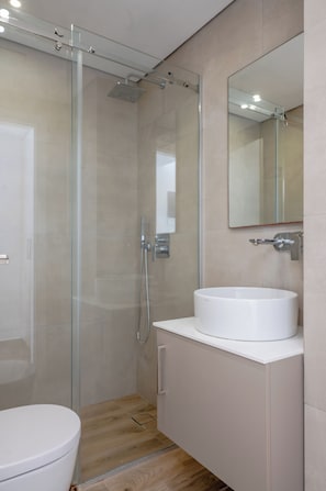 A modern bathroom with a glass-enclosed shower, a white sink, and a mirror above it. - Apeiron Perissa – Apeiron Hotels (Perissa)