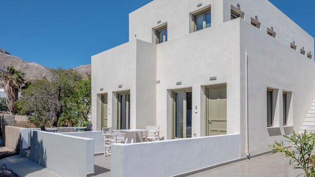 Un edificio moderno e bianco con molte finestre e porte, zona con posti a sedere all'aperto con tavoli e sedie bianche, e un cielo azzurro limpido sullo sfondo.