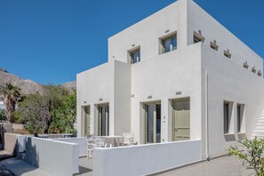 Een modern wit gebouw met meerdere ramen en deuren, een terras met witte tafels en stoelen, en een helderblauwe lucht op de achtergrond.