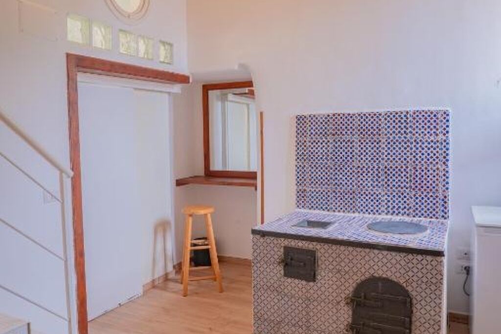 Uma cozinha compacta com painel decorativo, um banquinho de madeira pequeno e uma janela sobre a pia.