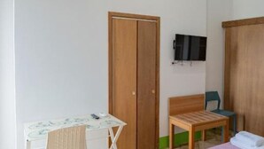 Ett litet rum med en trägarderob, en tv som är monterad på väggen, ett litet bord med en stol och en säng med en grön vägg.
