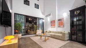 Una sala de estar moderna con una mesa de centro de vidrio, un sofá color beige, una mesita de noche de madera con una lámpara y una planta grande en maceta.