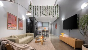 Una sala de estar moderna con sofá, mesa de centro y televisor. Se ve una cocina al fondo con taburetes de bar.