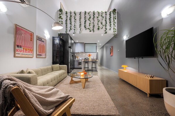 Una sala de estar moderna con sofá, mesa de centro y televisor. Se ve una cocina al fondo con taburetes de bar.
