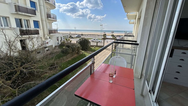 Ein Balkon mit rotem Tisch, mit Blick auf einen Strand und angrenzende Gebäude.
