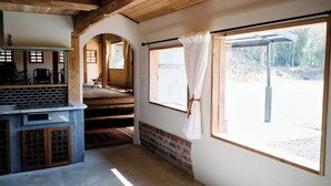 Una habitación con chimenea de ladrillo, vigas de madera en el techo, un ventanal con cortinas y vistas a un área boscosa.