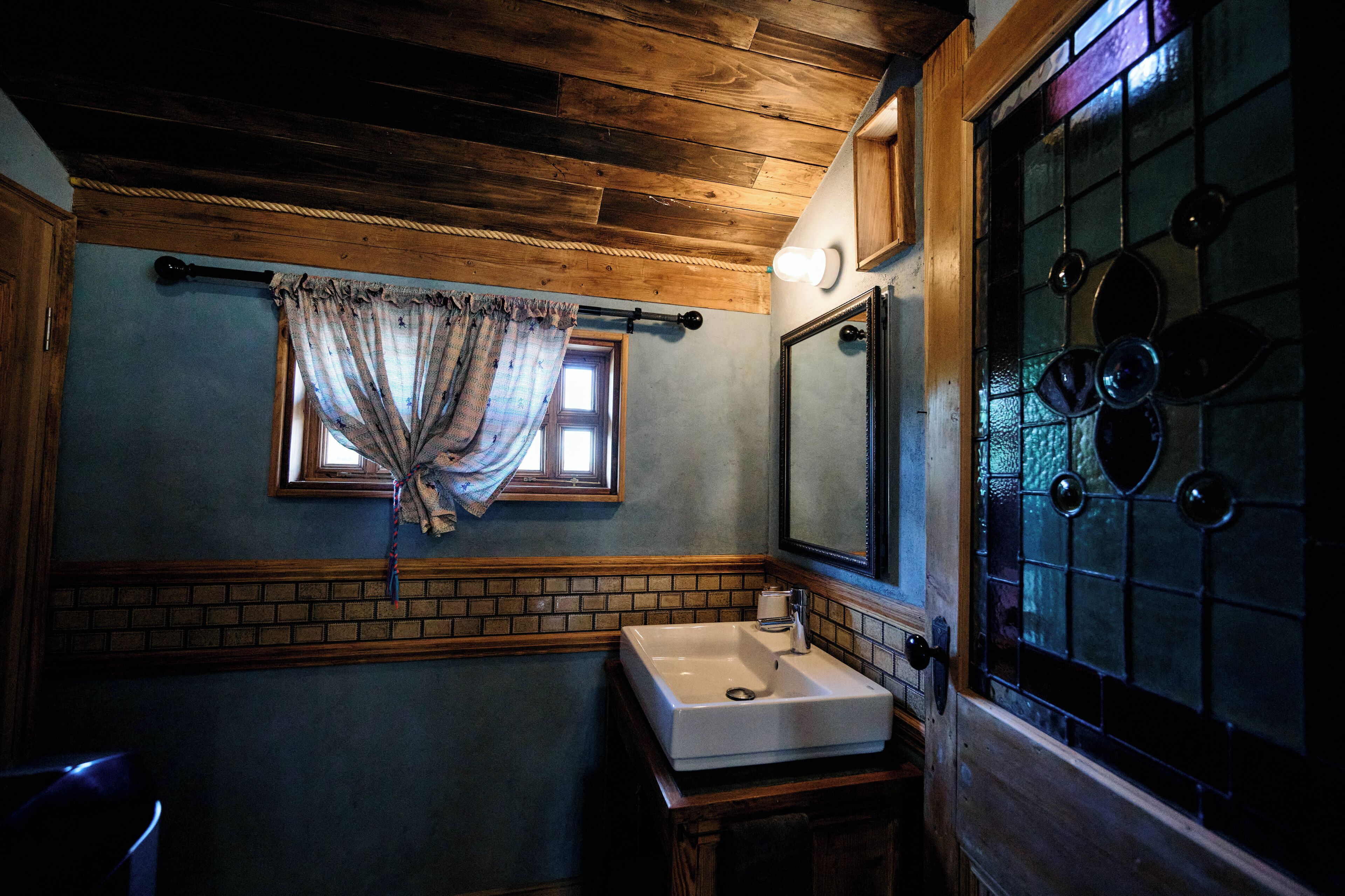 A bathroom with a wooden ceiling, a window with curtains, a sink, and a mirror.