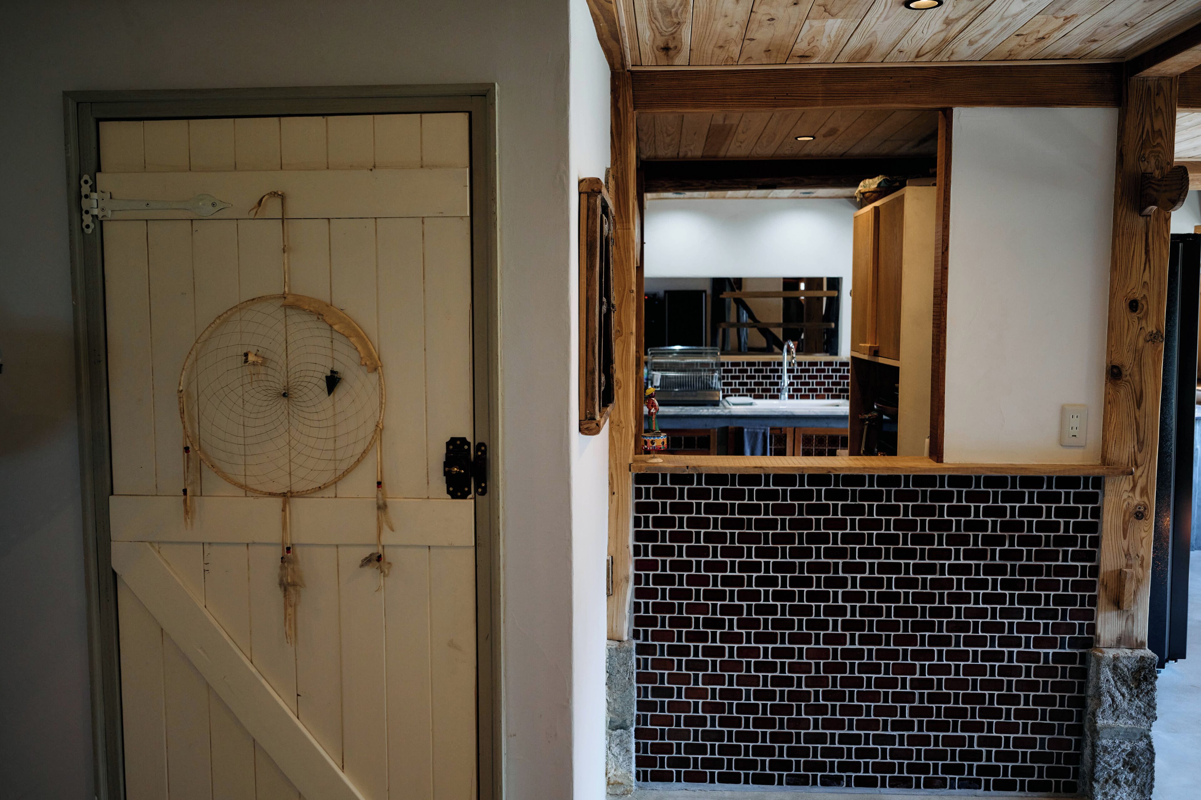 A wooden door with a dreamcatcher, a brick wall, and a glimpse of a kitchen area.