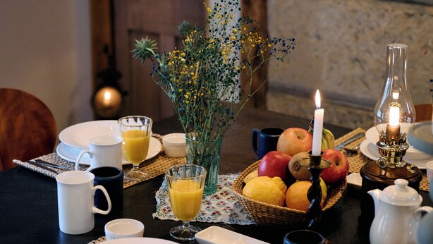 Una mesa de desayuno dispuesta con platos, tazas, un florero con flores, una vela y una cesta de frutas.