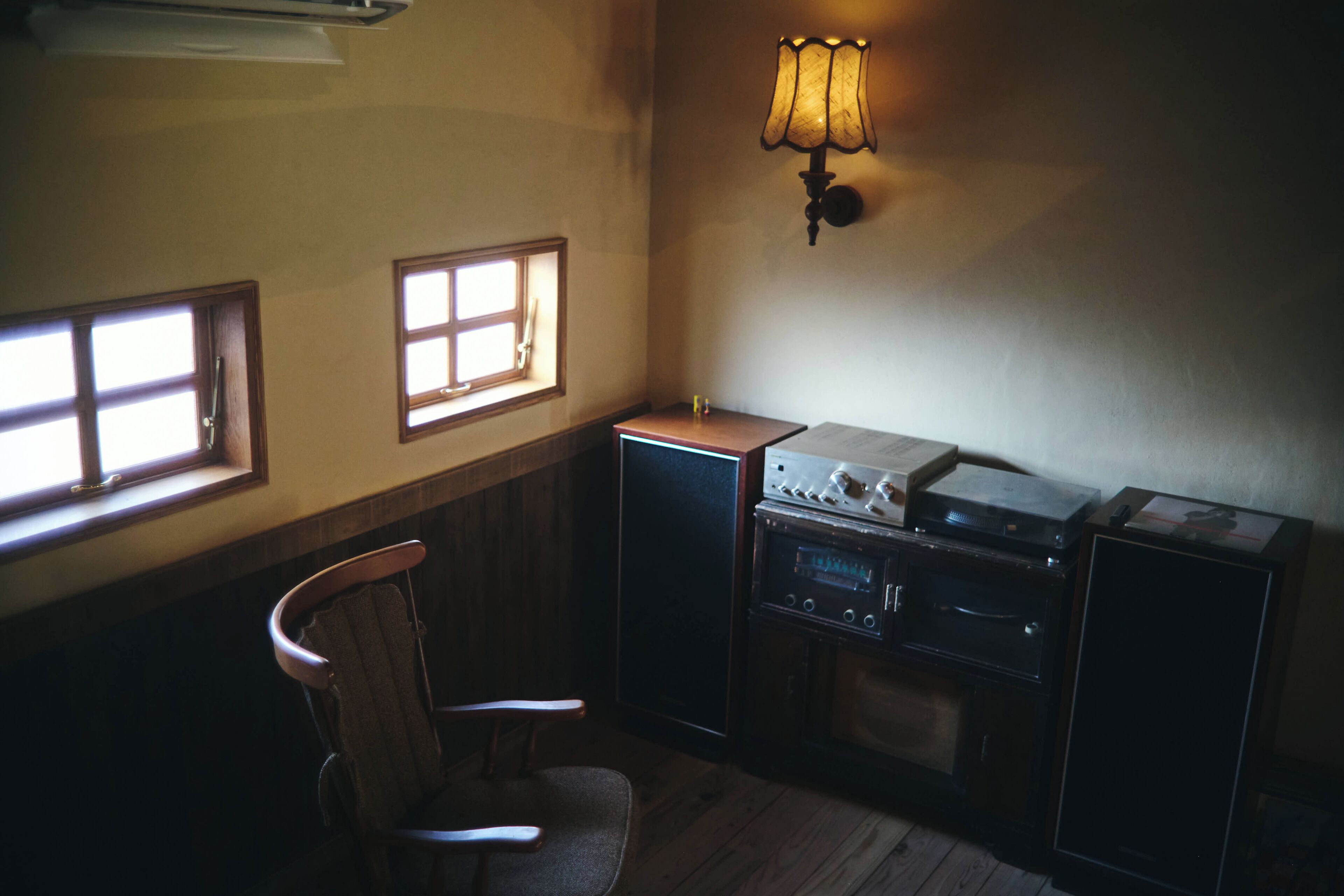 A room with a wooden chair, a small table, a wall-mounted lamp, and a window with a view.