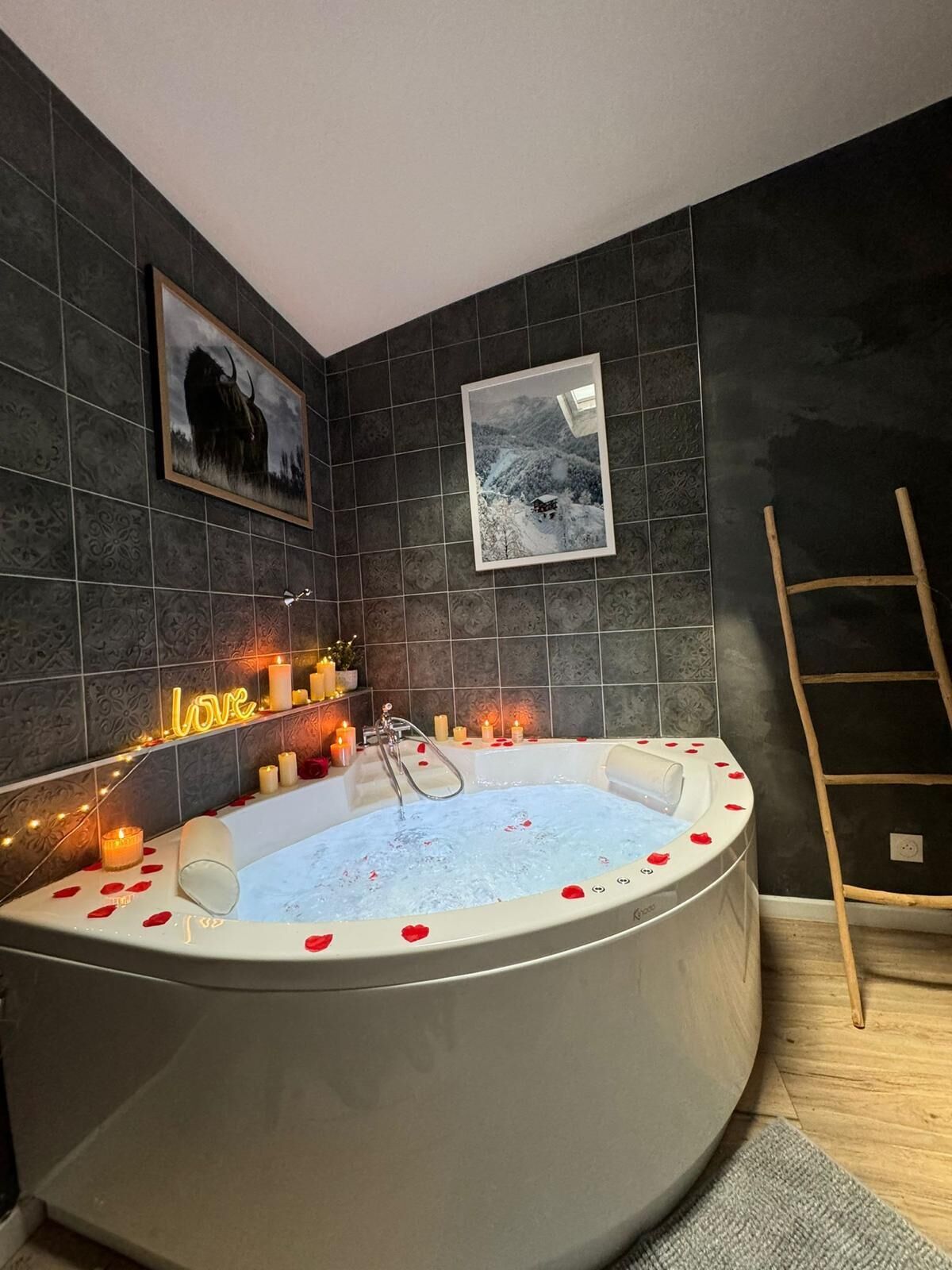 Ein modernes Badezimmer mit einer großen Badewanne, dunklen Fliesenwänden, gerahmten Bildern, Kerzen und einem neonbeleuchteten 'love'-Schild.