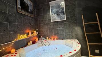 Ein modernes Badezimmer mit einer großen Badewanne, dunklen Fliesenwänden, gerahmten Bildern, Kerzen und einem neonbeleuchteten 'love'-Schild.